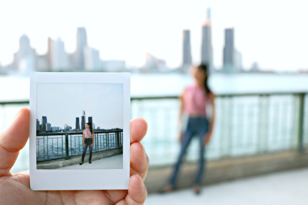 An Instax square print is held up to the camera with a girl and the Detroit skyline in the background. 