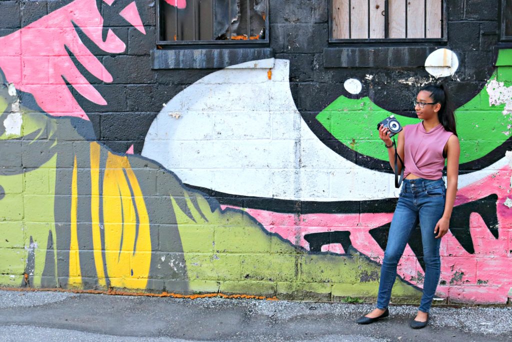 A girl stands in front of a graffiti wall holding an Instax Square SQ6 camera.