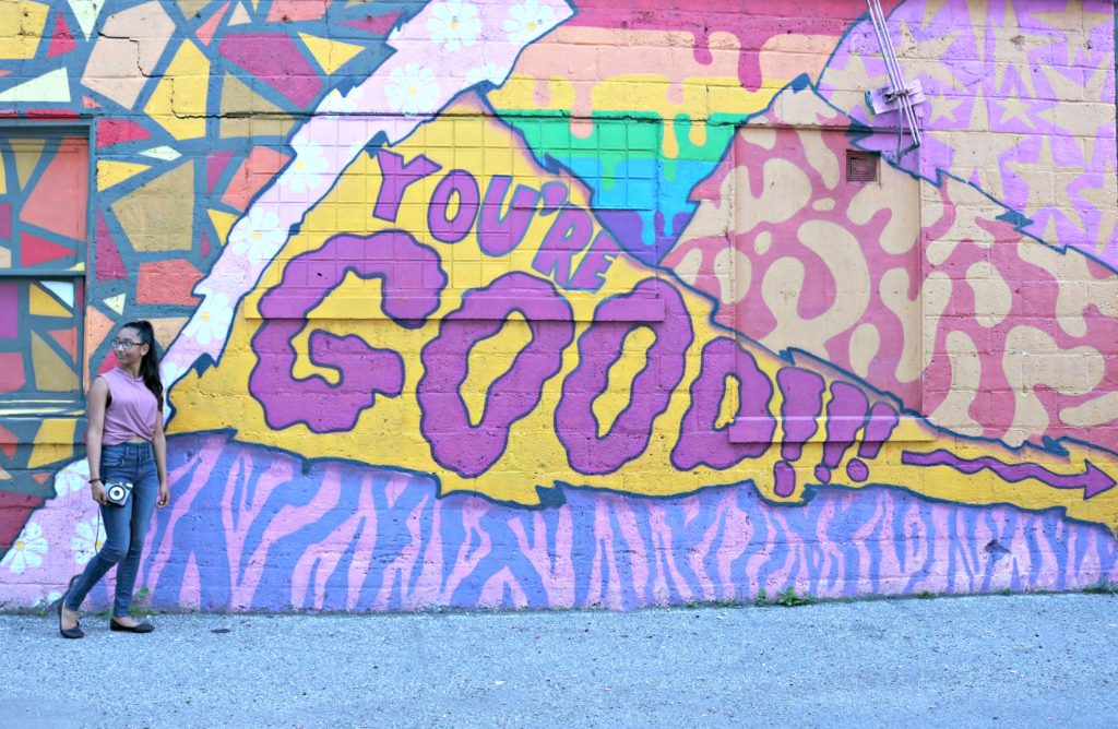 A girl stands in front of huge wall of graffiti with her Instax camera in hand.