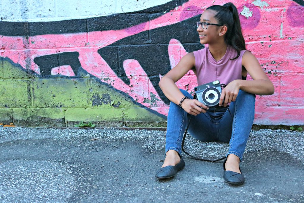 Girl sits in front of a wall and takes a break from taking pictures. 