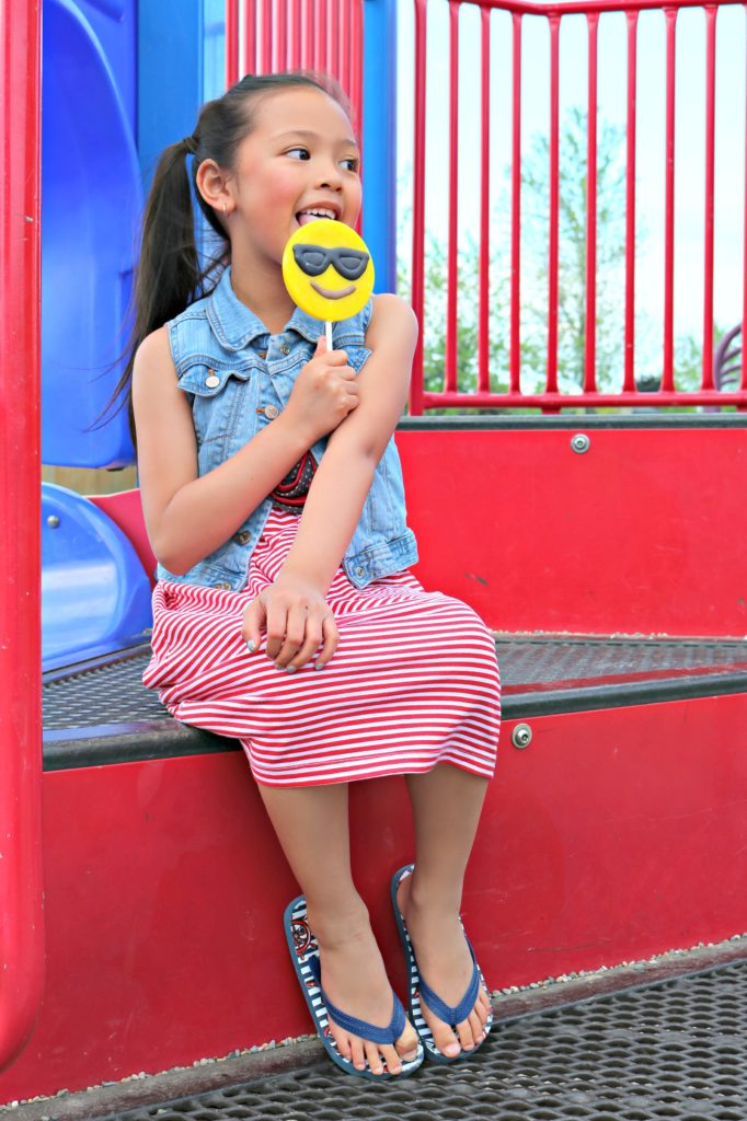 A little girl sits in the jungle gym and takes a break with an emoji lollipop. 