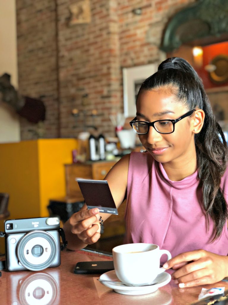 A girl sips on a coffee and looks at an Instax square print she just took.