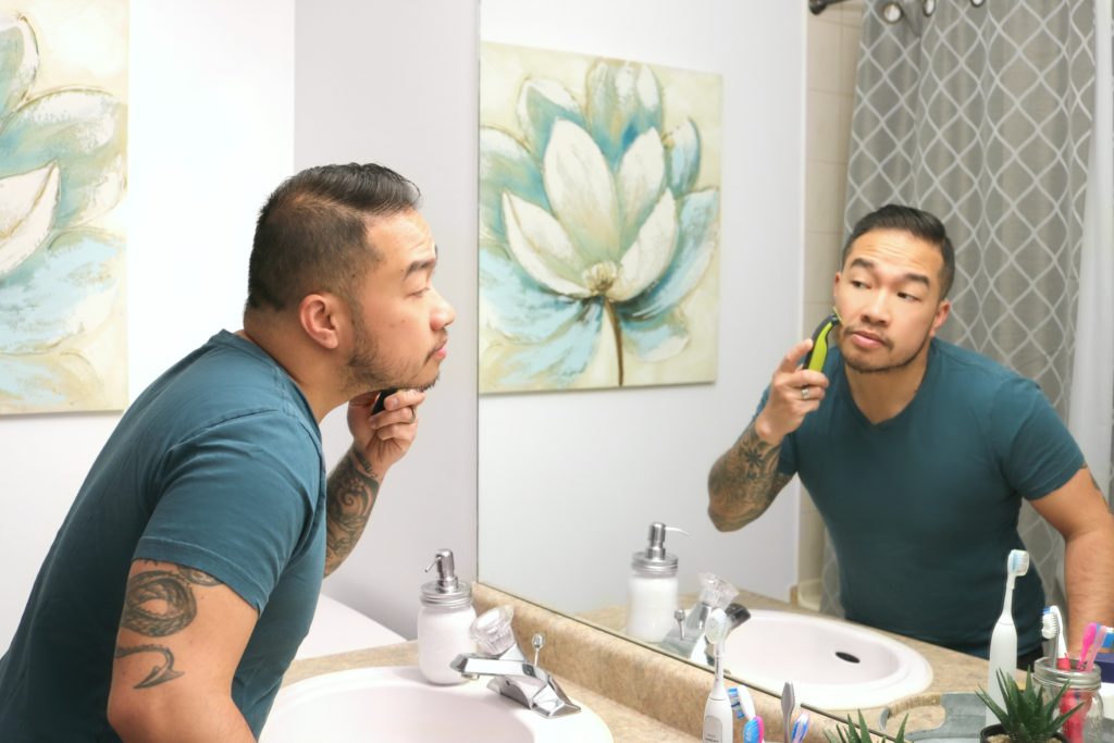 A man trips his beard in the reflection of the mirror. 