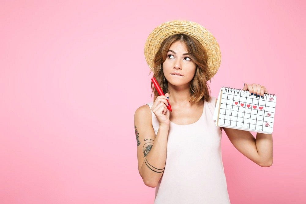 A woman holds a marker while marking her menstrual cycle on a calendar, she looks away in thought.