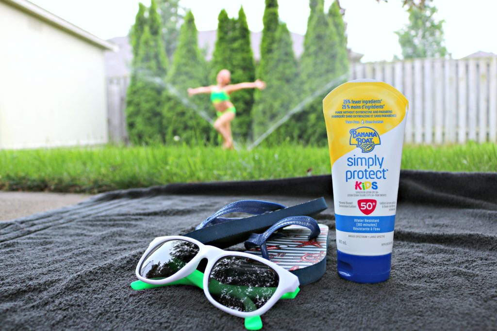 A little girl plays in a sprinkler in the background while sun screen, glasses, and flip flops on top of a towel are shown.