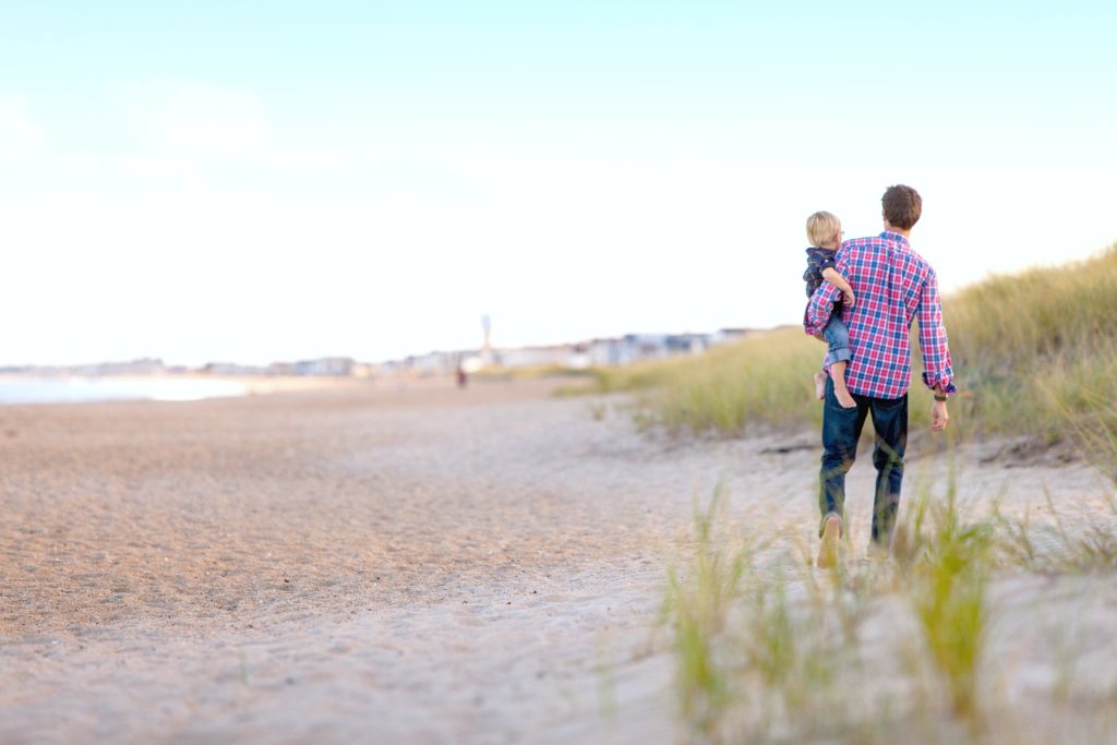 A father holds his son on his hip walking on an empty beach. 