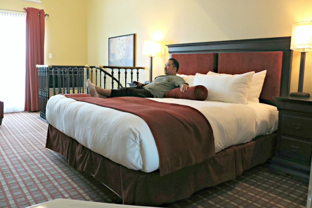 A man relaxes on a king-size bed and watches TV.