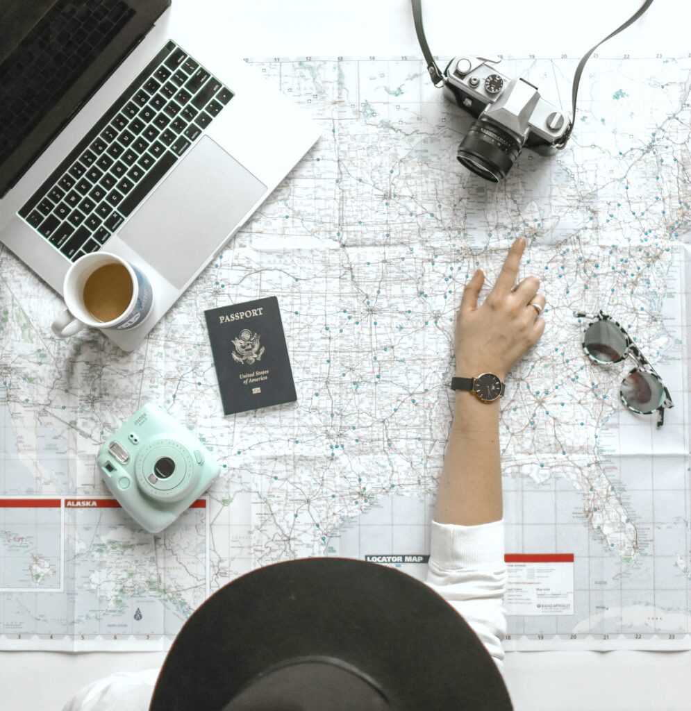 A woman plans her next trip and points to a location on a map. A computer, camera, passport, and glasses are shown.