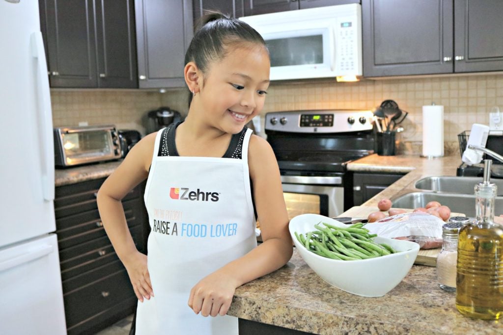 A little girl wears a Zehrs Take the Pledge - Raise a Food Lover apron and looks at her counter full of ingredients. 