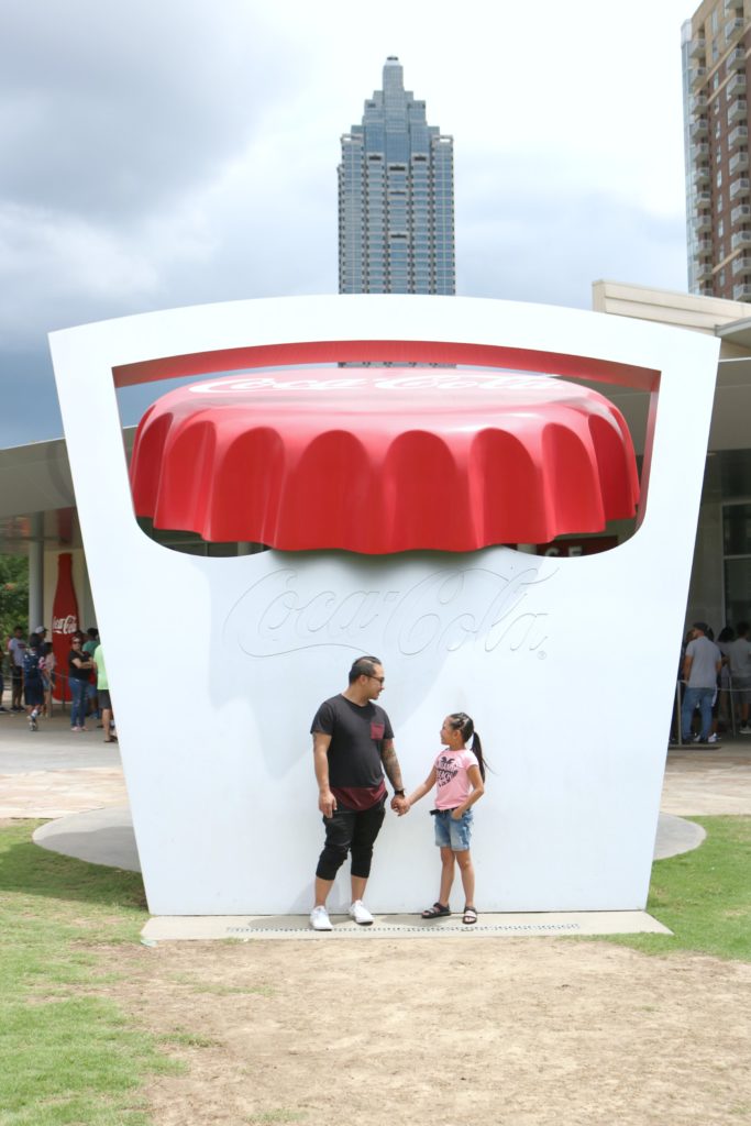 A little girl and her dad hold hands in front of the iconic bottle cap opener at World of Coca-Cola.