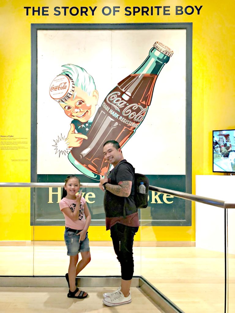 A father and his daughter stand in front of Sprite Boy sign at World of Coca-Cola.