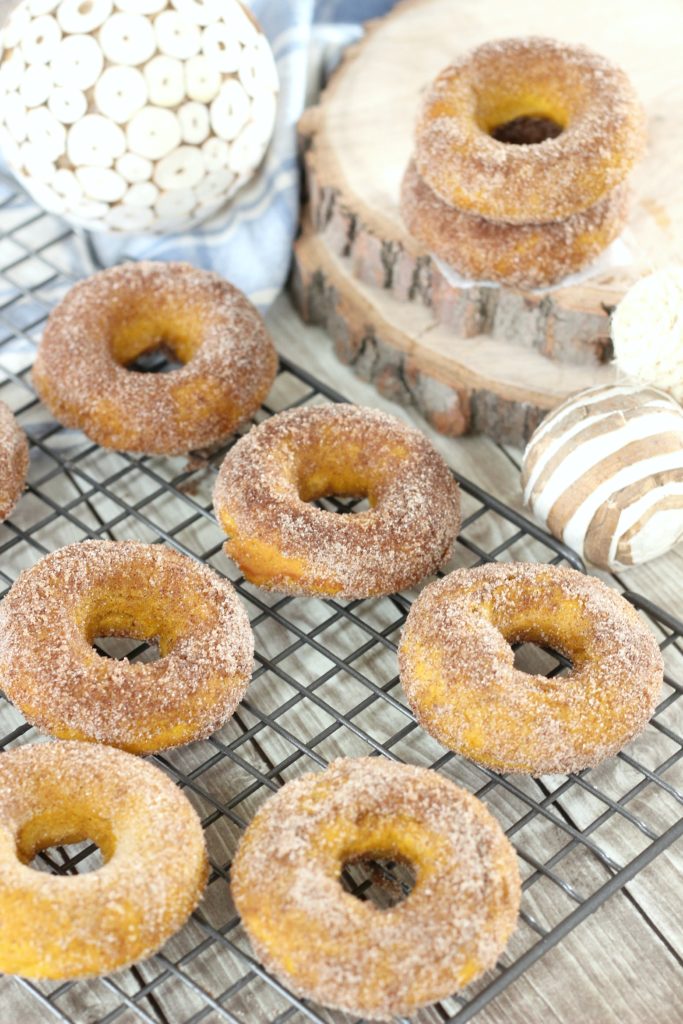 Baked pumpkin spice donuts are shown on a cooling rack with fall decor surrounding them.