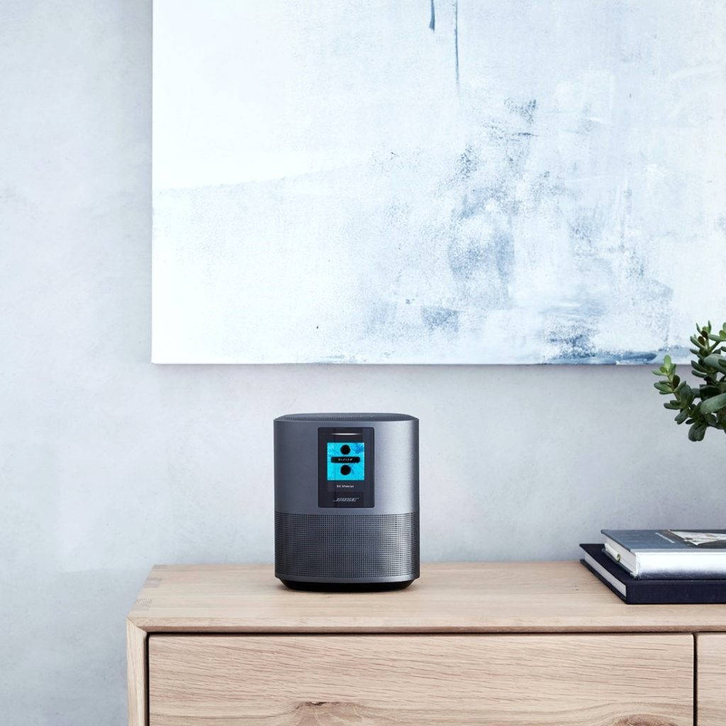 A slick Bose speaker sits on table with a painting hung on a wall behind it.