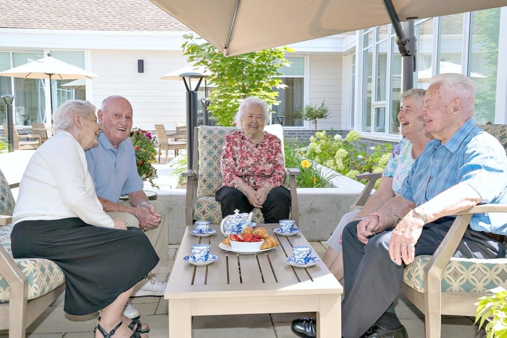 5 elderly people sit and enjoy tea and croissants on a patio.