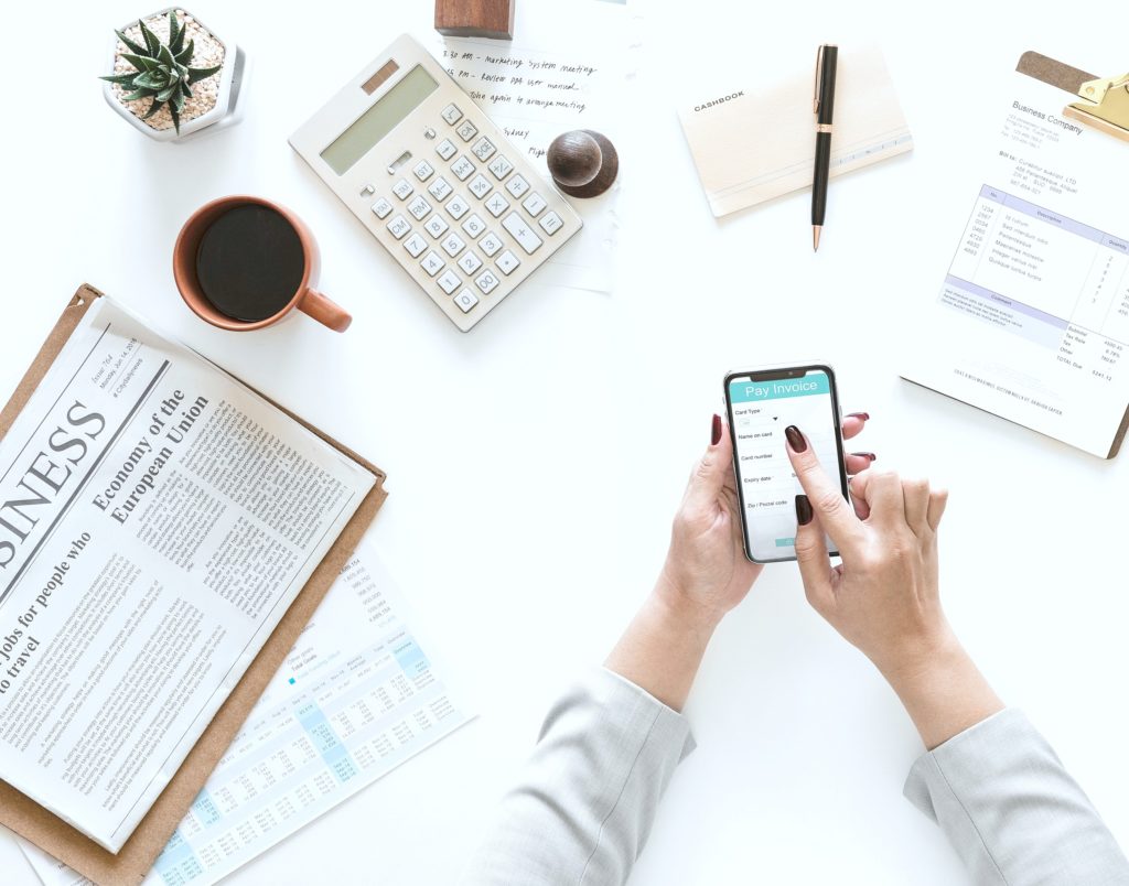 Woman pays invoice on her mobile phone while drinking coffee and balancing her books.