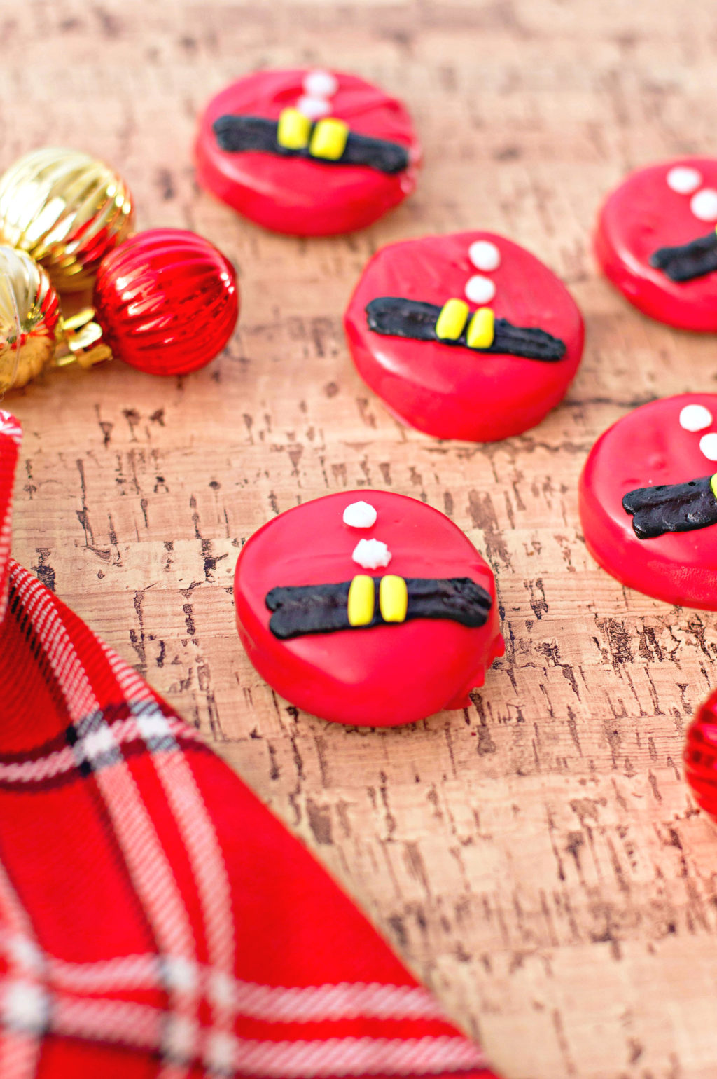 Santa Belt Chocolate Covered Oreos are shown against a cork background with a plaid napkin and mini gold and red ornaments. 