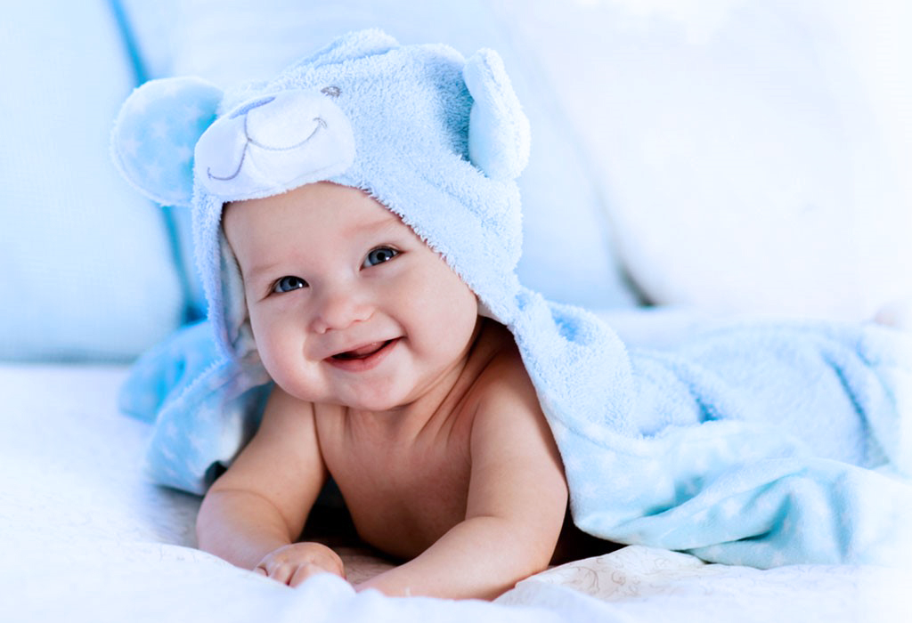 A baby smiles with a blue teddy bear towel on his head and body.