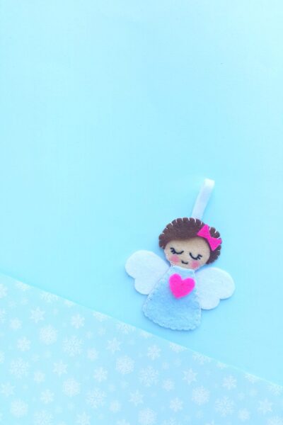 A felt angel Christmas ornament is laying on a blue background with white snowflakes.