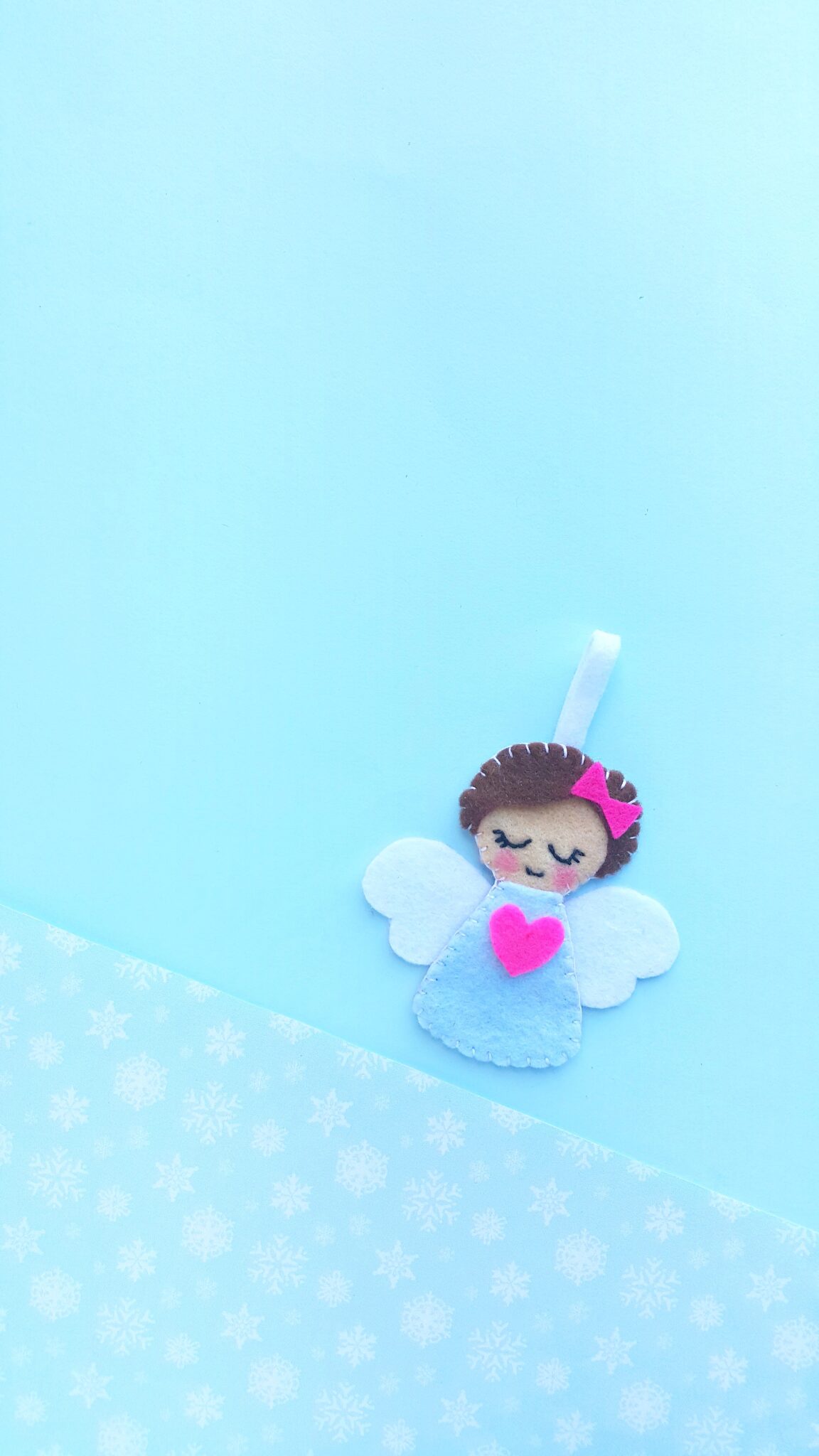 Felt angel with pink heart on dress, brown hair with a pink bow against a blue background.