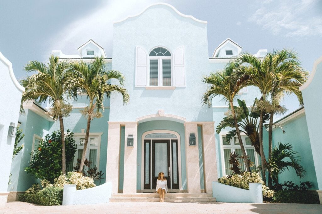 Woman sits in the front step of her large pastel blue and white house. 