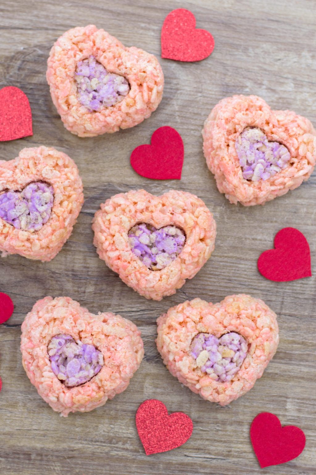 Valentine's rice cereal treats in shape of hearts on wood background with red hearts surrounding them.