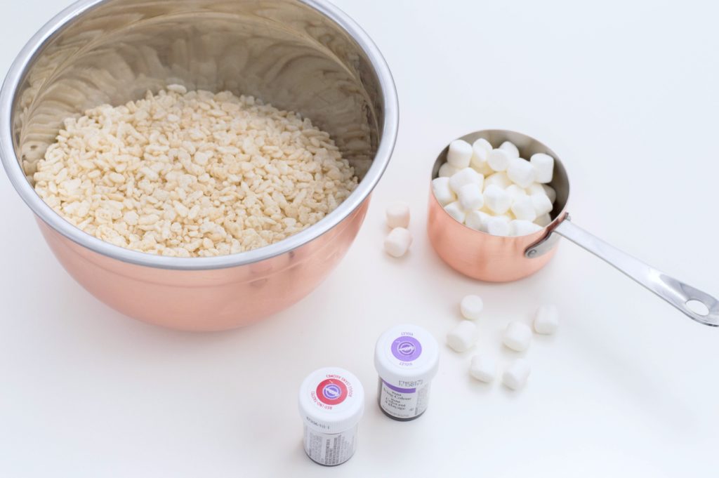 A rose bowl mixing bowl is filled with rice cereal, a measuring cup is filled with marshmallows, and pink and purple food coloring is shown. 