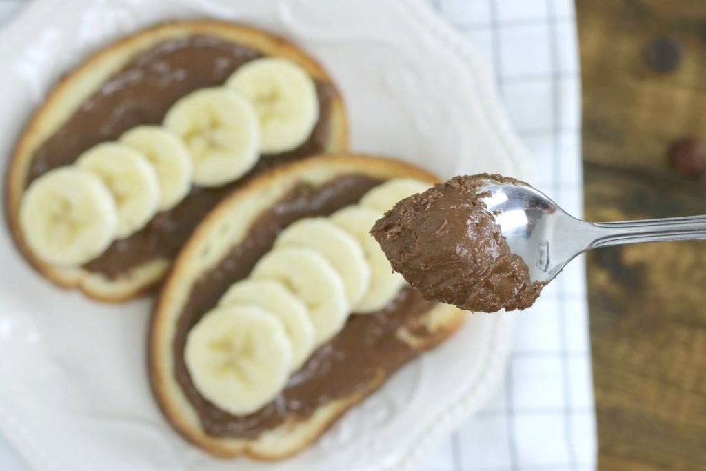 Toast with hazelnut spread and banana with spoon showing a spoonful, while toast is out of focus on a plate on top of a napkin.