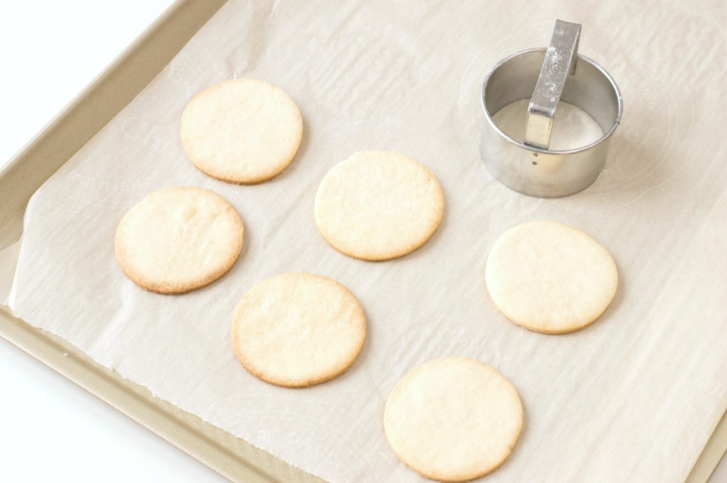 A baking sheet lined with parchment paper holds 6 sugar cookies and a cookie cutter. 
