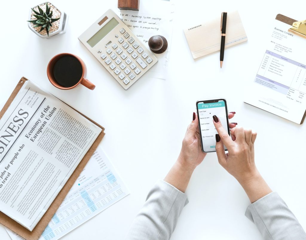 Flatlay view of woman using her smartphone to pay bills. She is surrounded by a calculator, newspaper, pen, paper, coffee, an invoice and a succulent plant.