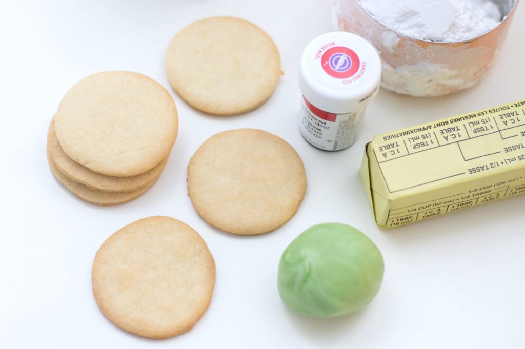 Six sugar cookies that are unfrosted, with a stick of butter, green fondant, red food colouring, and powdered sugar are the ingredients needed. 