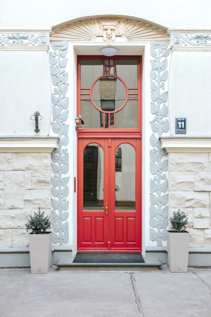 A grey and white frame with a bright red door, with two plants lining each side.