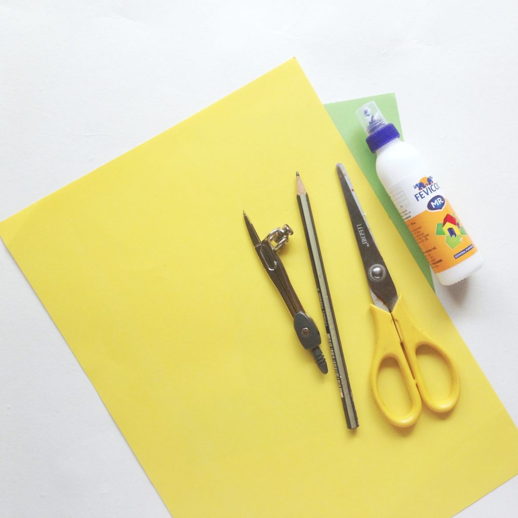 Supplies needed to make paper flowers, glue, scissors, construction paper, compass, pencil are shown.