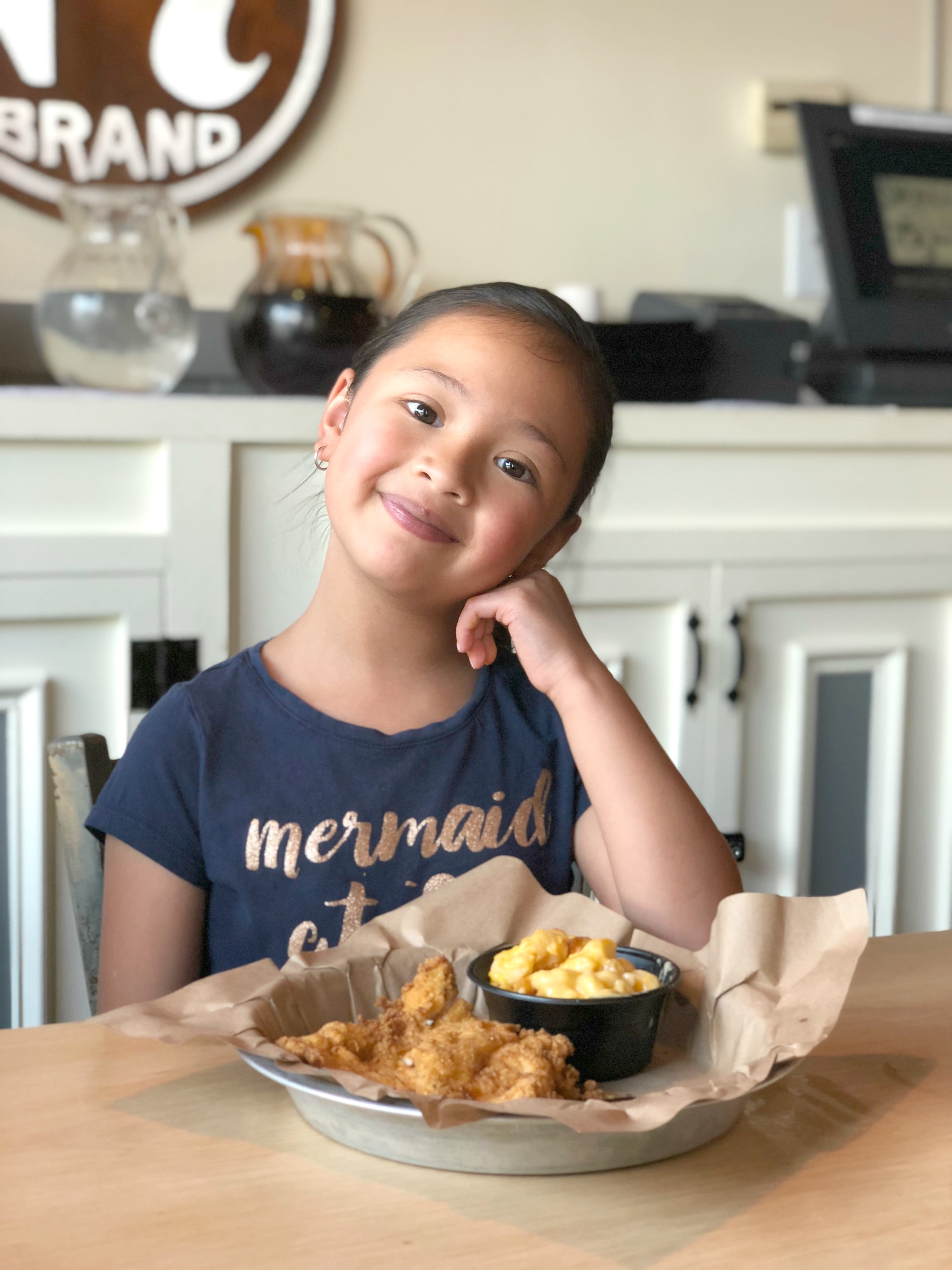 Little girl sits inside Smoke Jack in Alpharetta, GA with a tray of chicken fingers and mac and cheese, she smiles. 