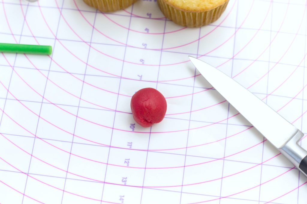 Red fondant ball on a Wilton measuring chart, knife, cupcakes, and brush is pictured.
