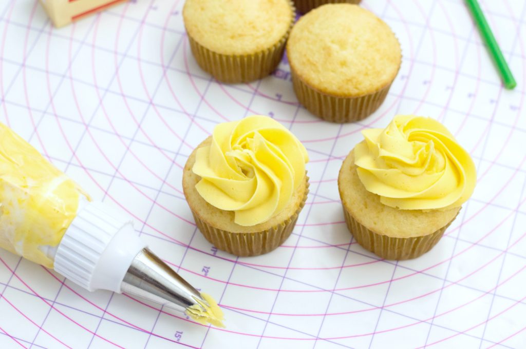 Cupcakes being frosted with yellow frosting, cupcakes and piping bag is shown.