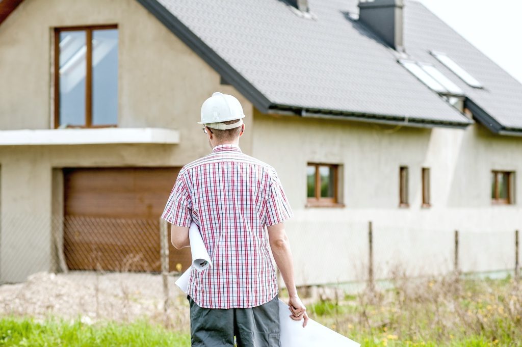 Man with hard hat on is holding building plans and standing in front of a house that is under construction. 