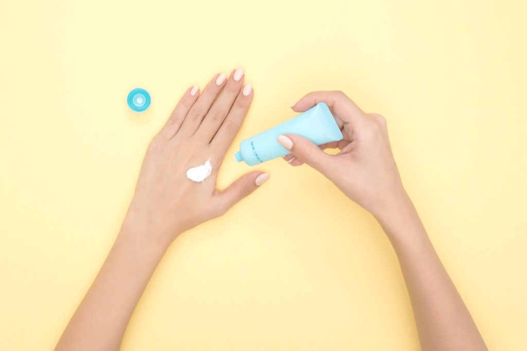 Woman applies sunscreen onto her left hand on top of a bright yellow background. 