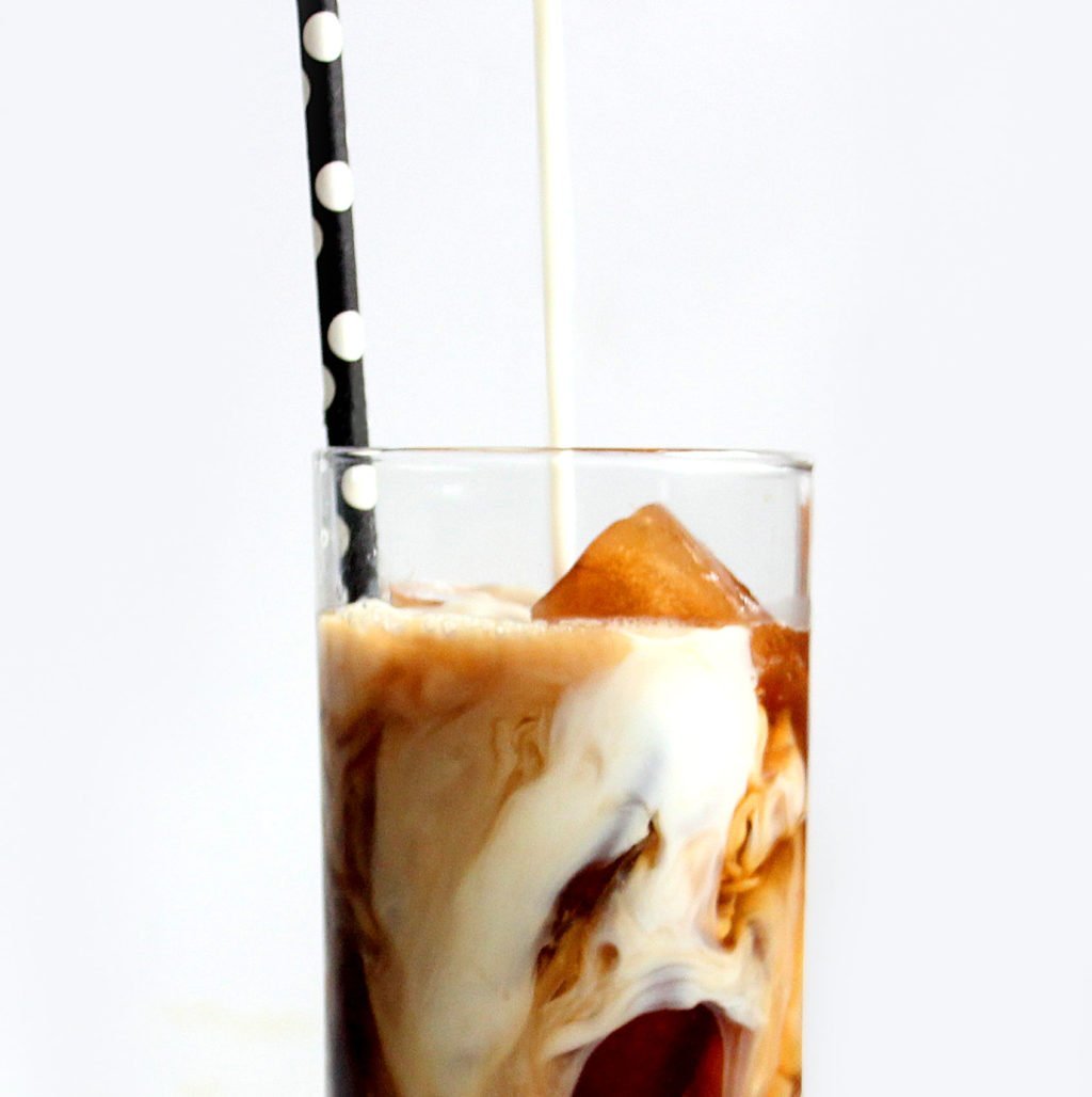 Milk being poured into iced coffee in a clear glass with a polka dot straw.