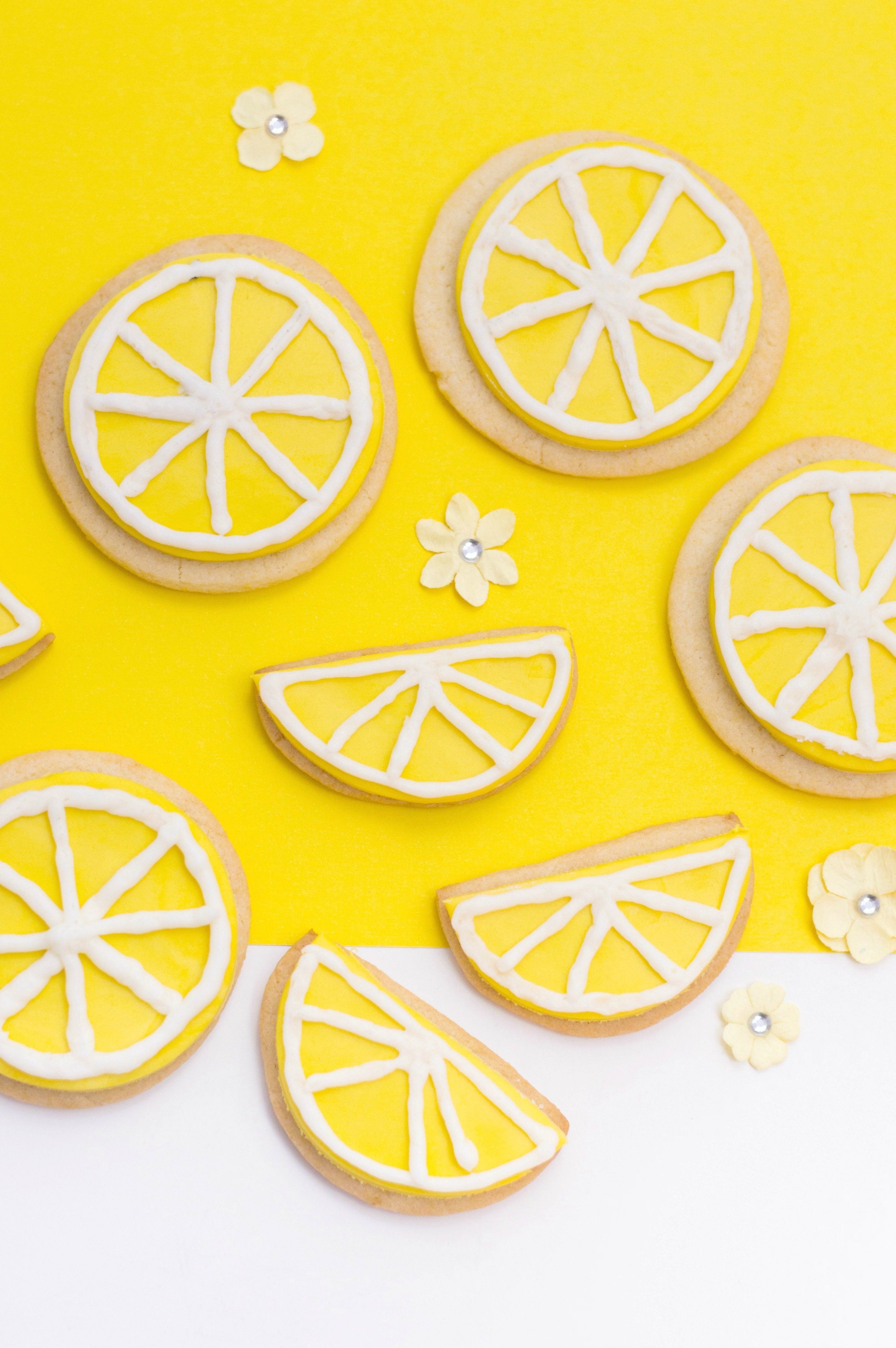 A yellow and white background with sugar cookies and small flowers are shown. 4 are made to look like lemon slices and 4 are cut in half to look like lemon wedges.