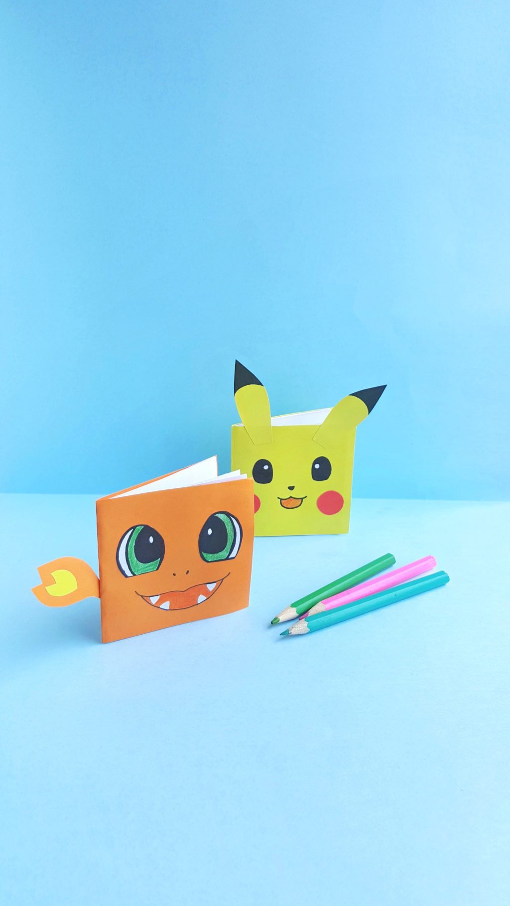 A mini Charmander notebook and Pikachu notebook are shown standing up with coloured pencils. 