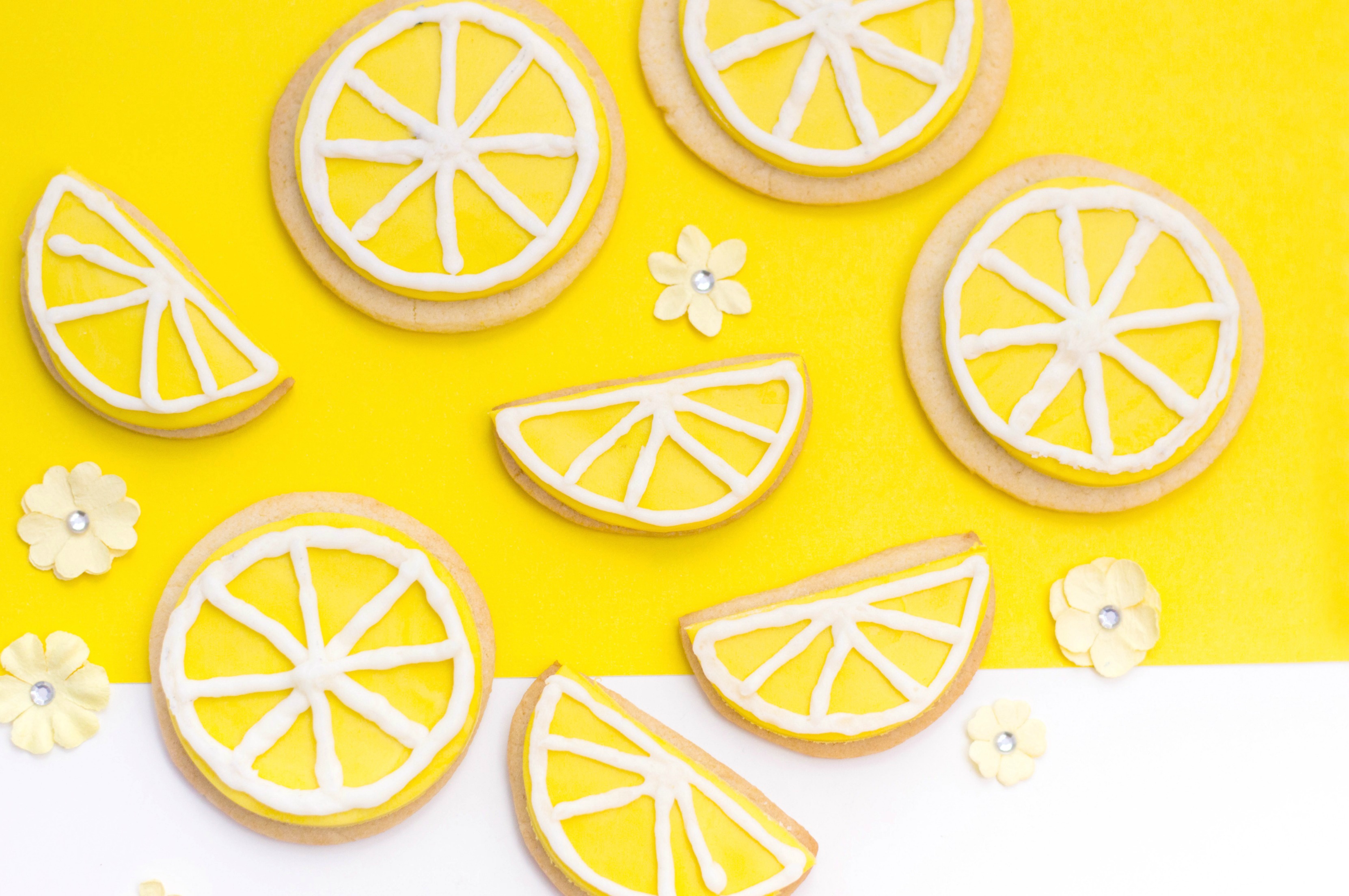 A yellow and white background with little yellow flowers, cookies and half cookie wedges placed on top. Iced and they look like lemon wedges and lemon slices ready to eat!