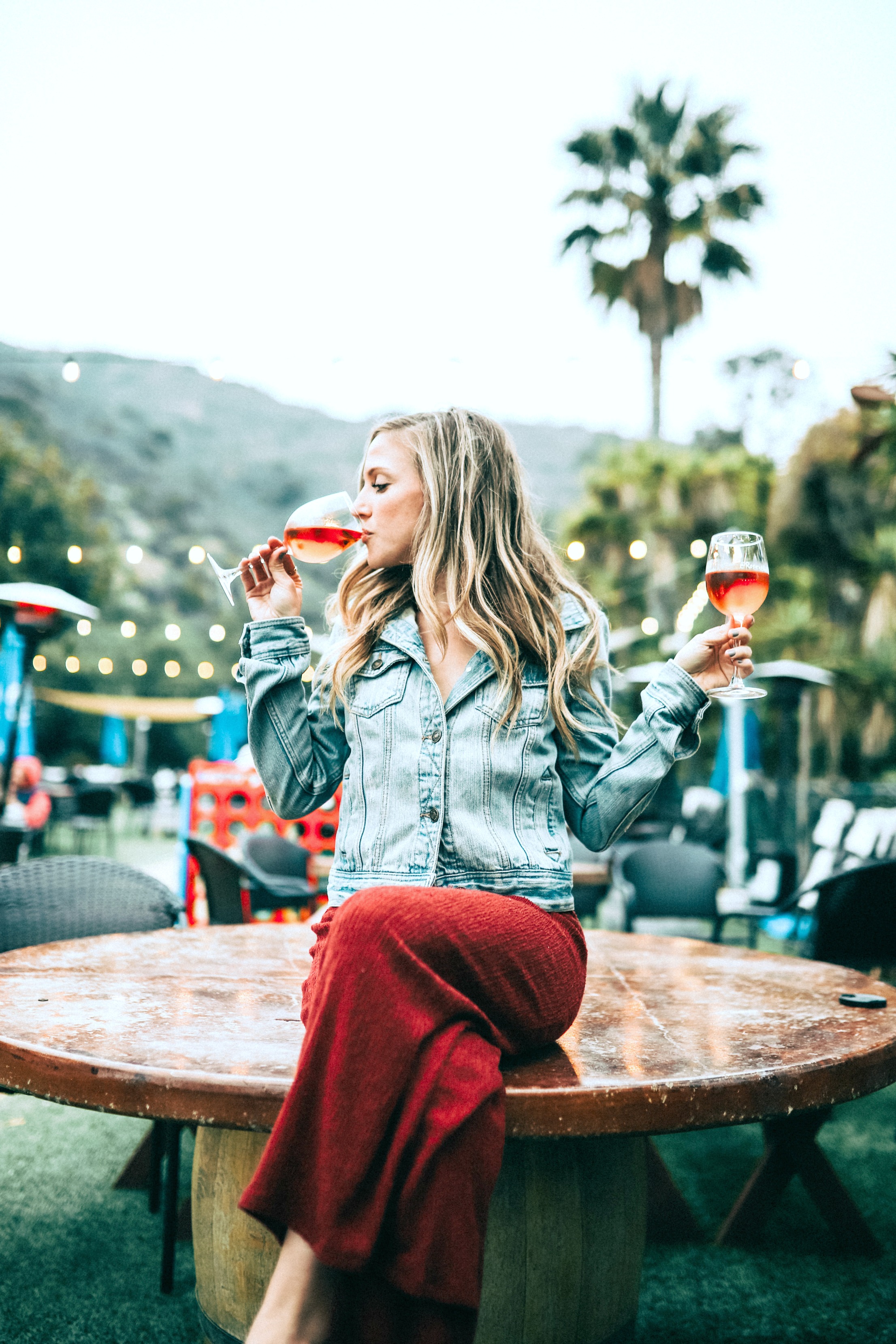 A woman sitting on a table drinking a glass of wine while she holds another glass of wine. 