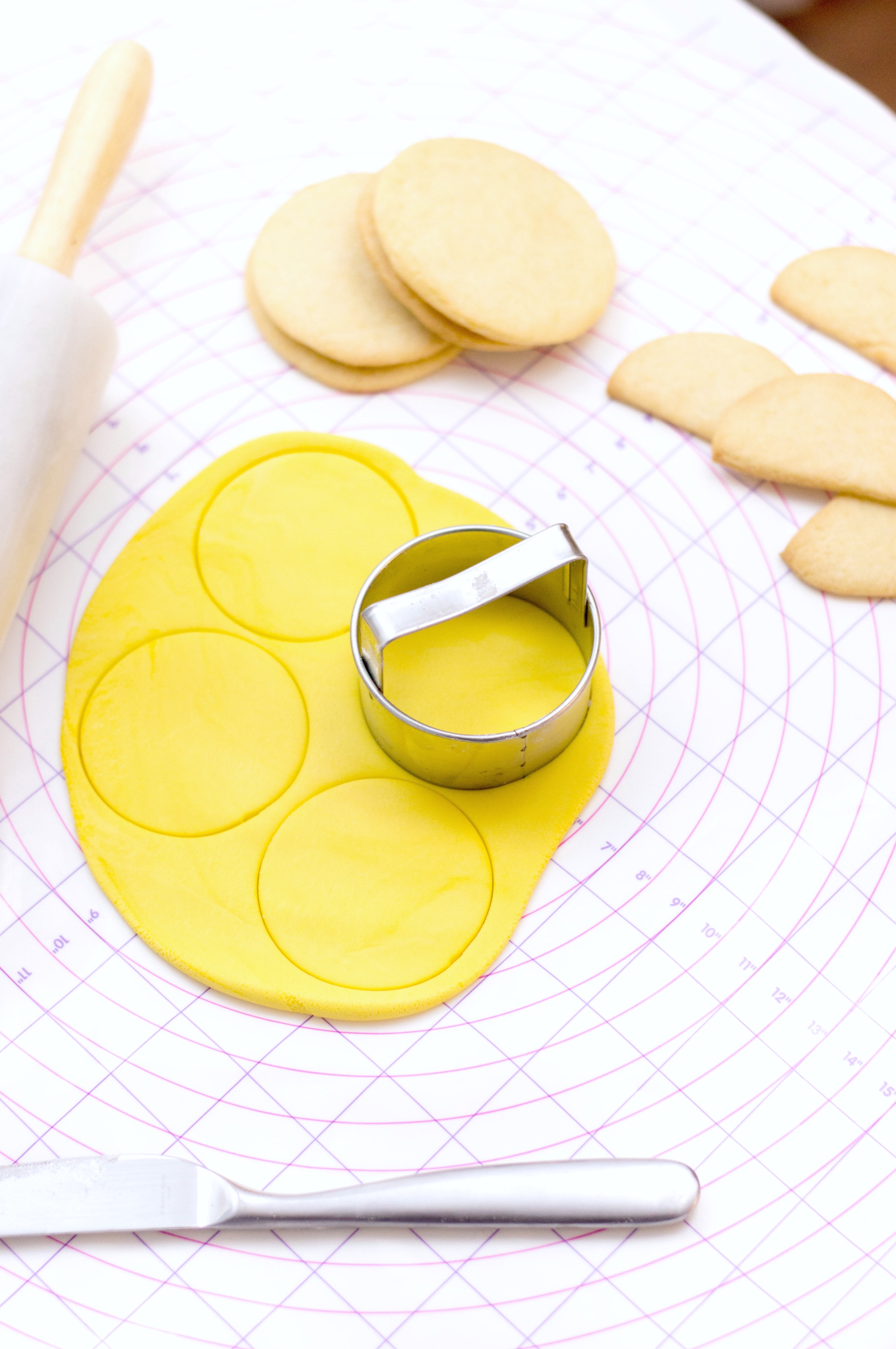 Yellow fondant is rolled out and cookies are piled up. Circles are being cut out with a cookie cutter to place on top of cookies. 