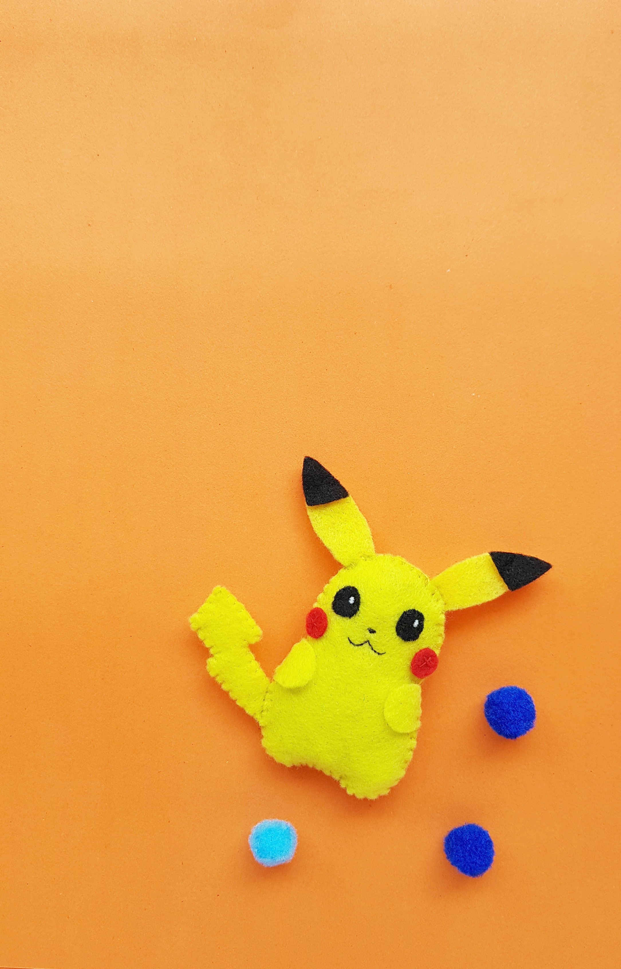 A cute felt Pikachu plushie craft is sewn together and sits on top of an orange background with 3 poms poms surrounding him. 