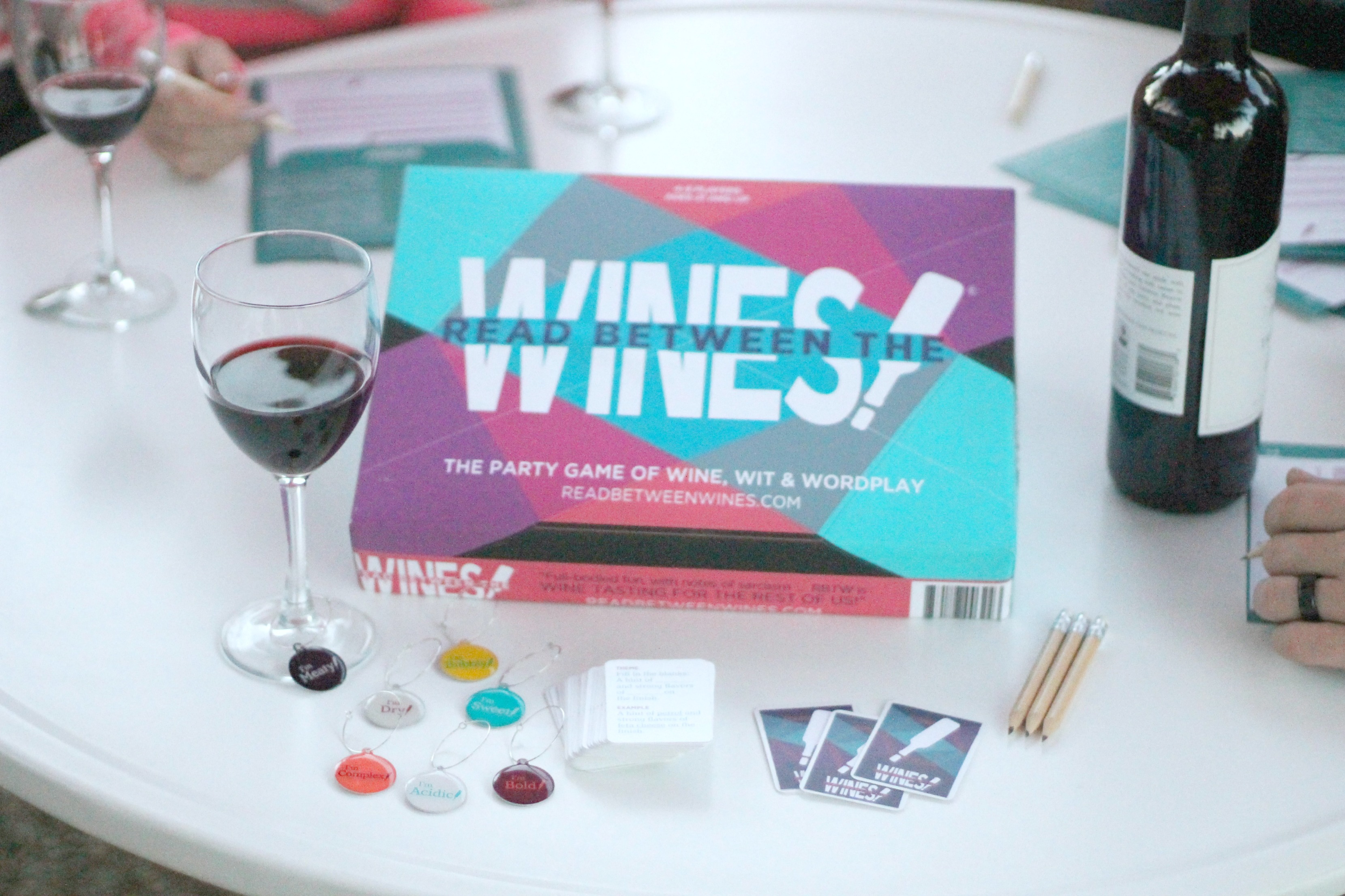 A fun board game, Read Between the Wines is shown on a table with glasses of wine and ladies gathered to play the board game. 