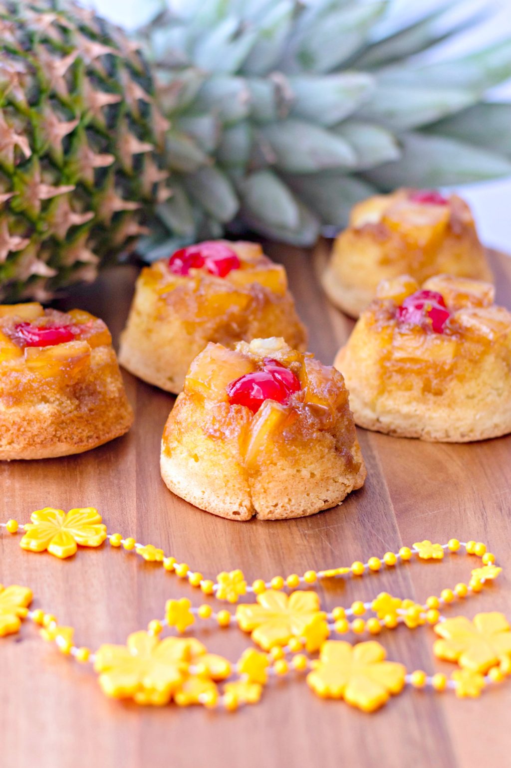 A pineapple lays on its side, with 5 mini pineapple upside down cakes and a yellow plastic lei.