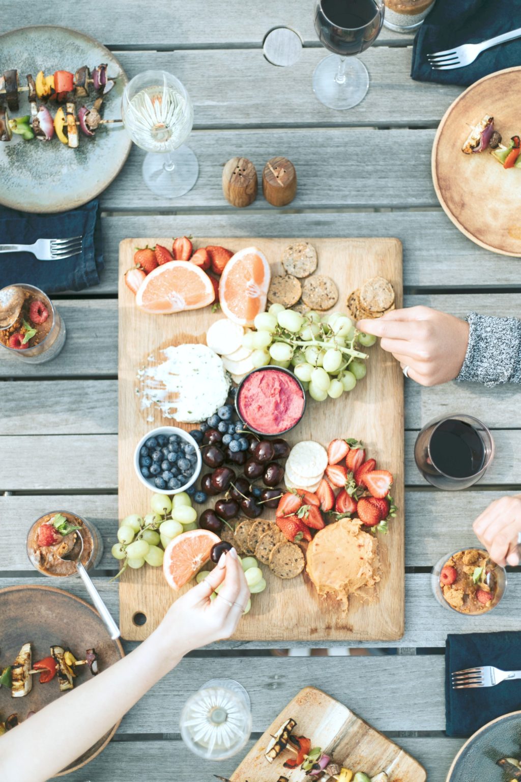 A flat lay of a charcuterie board with various hands taking food. Each plate has BBQ skewers on it. 