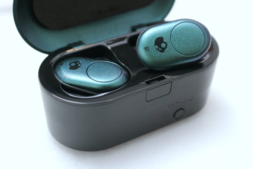 A charging case with the Skullcandy Push earbuds, one is plugged in, the other is showing the camera. 