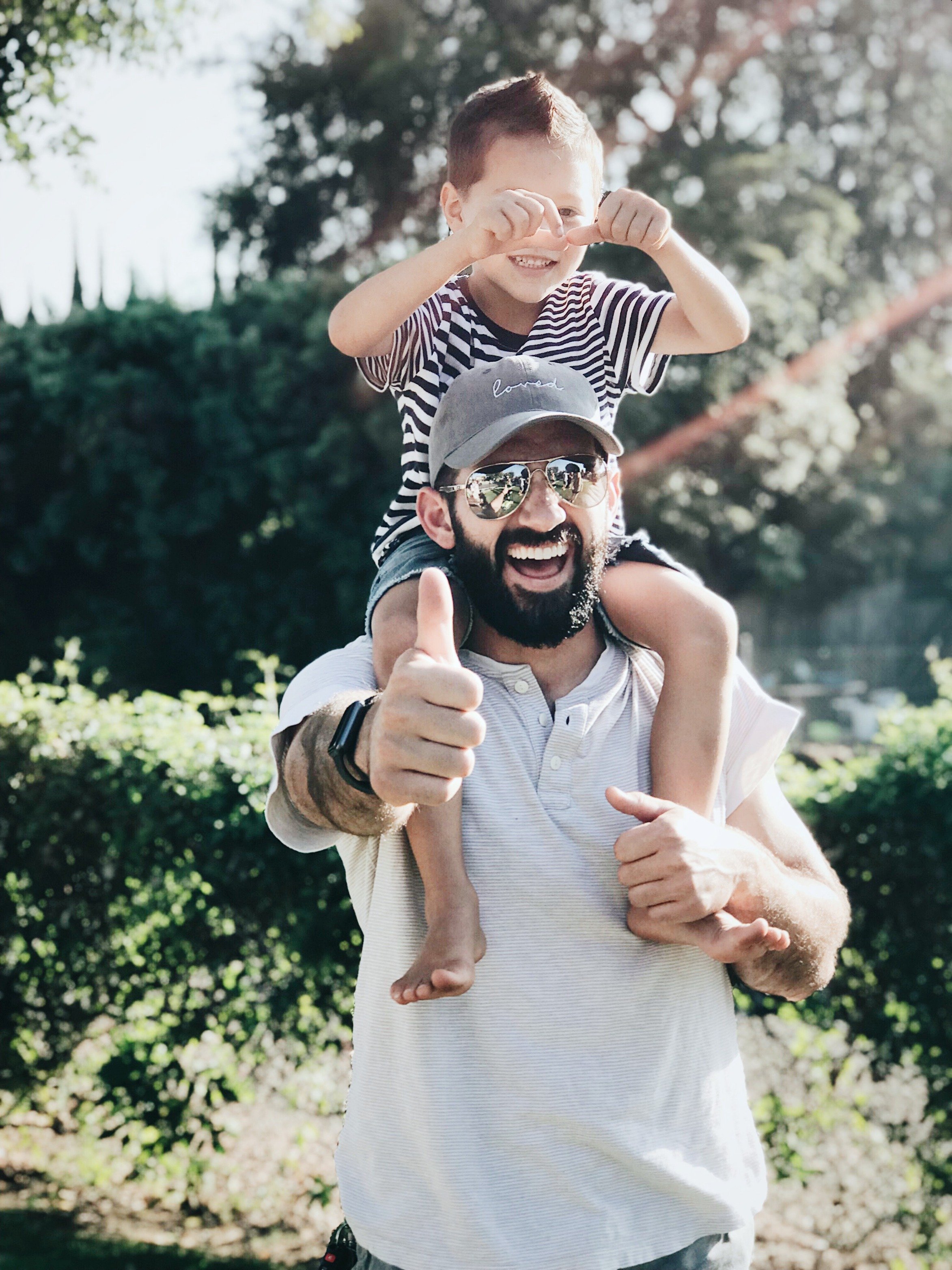 A dad wearing sunglasses holds his son on his shoulders, he smiles and gives the camera a thumbs up. Following is a list of the best experiences to give Dad.