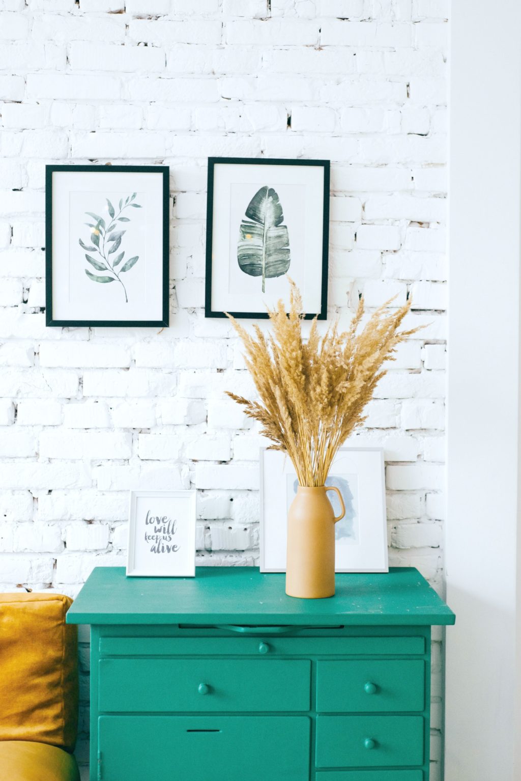 A painted white brick wall, with a teal nightstand with two pictures on the wall and flowers on the table. An easy way to decorate on a budget. 